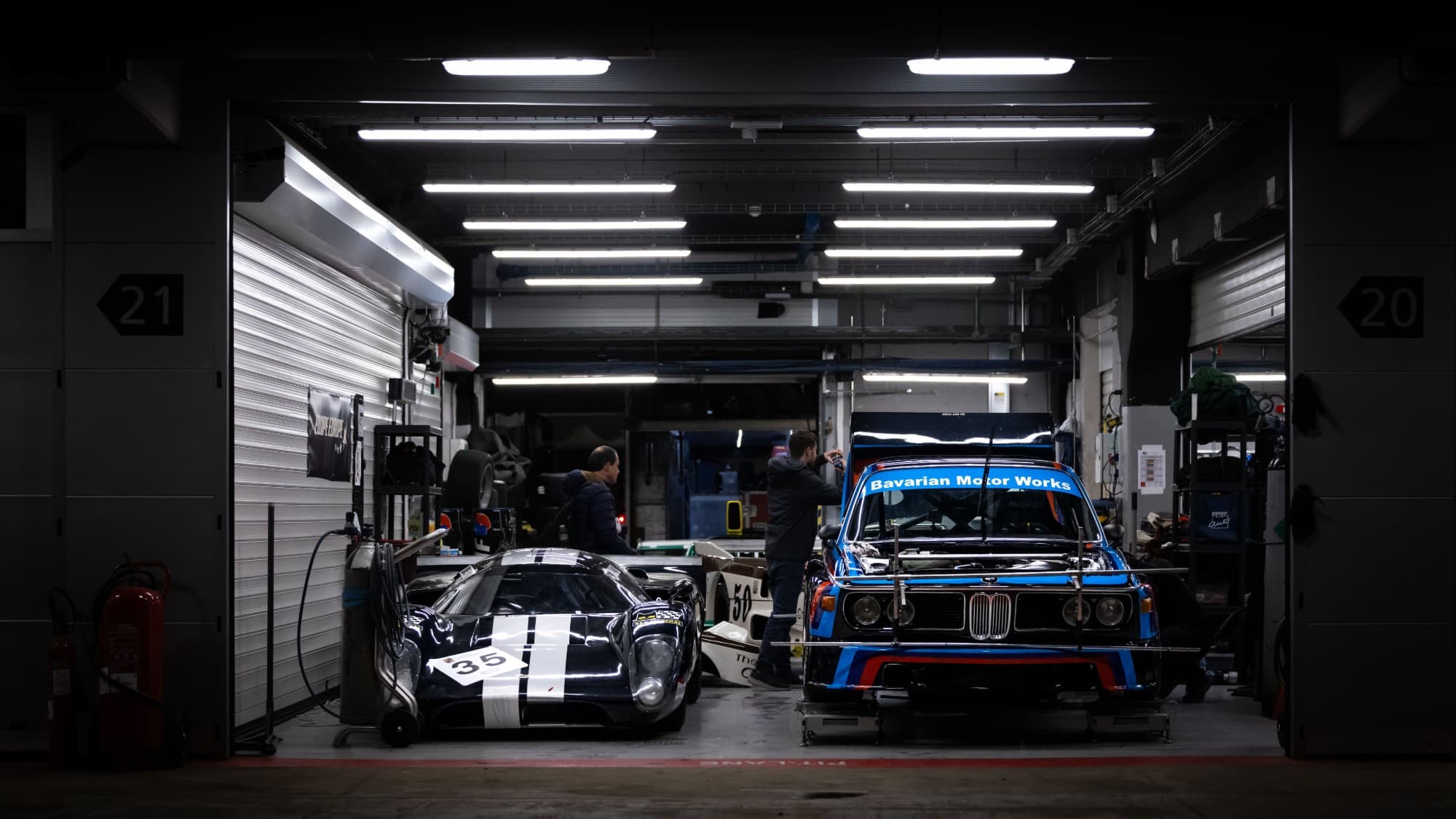 A vintage BMW 3.0 CSL race car and an endurance racer side by side in a night pit garage