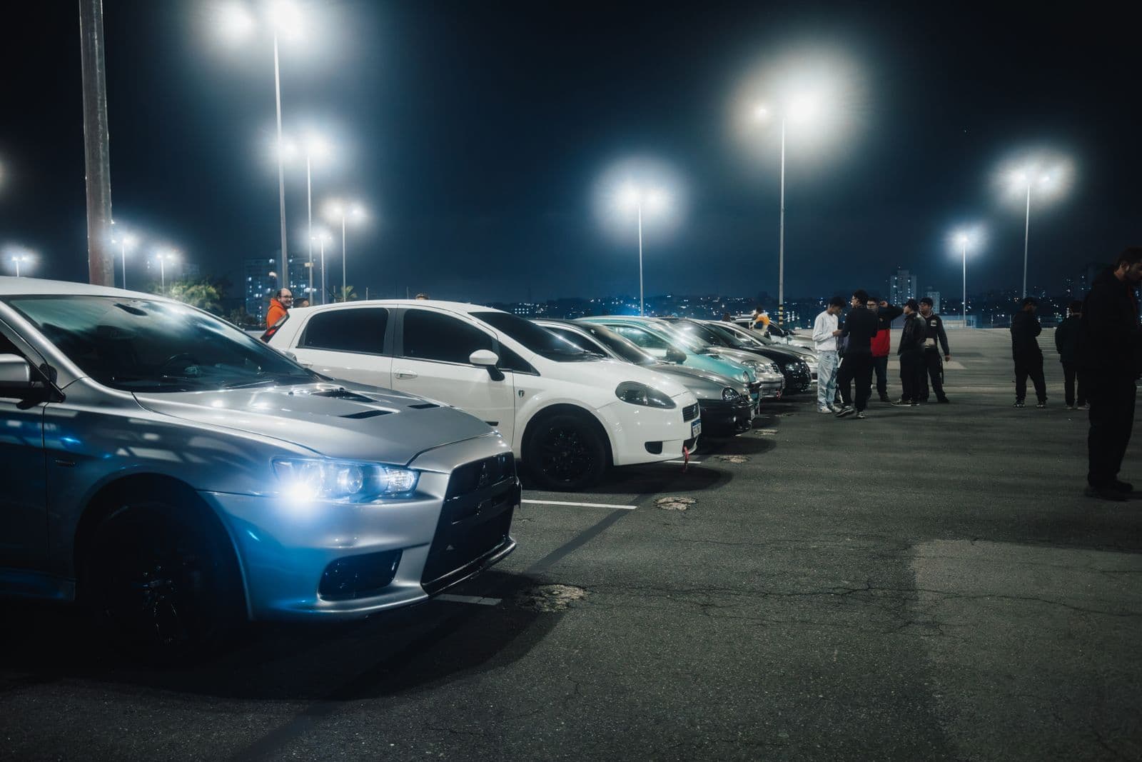 A row of modified cars at a night meet under parking-lot floodlights, owners talking between them