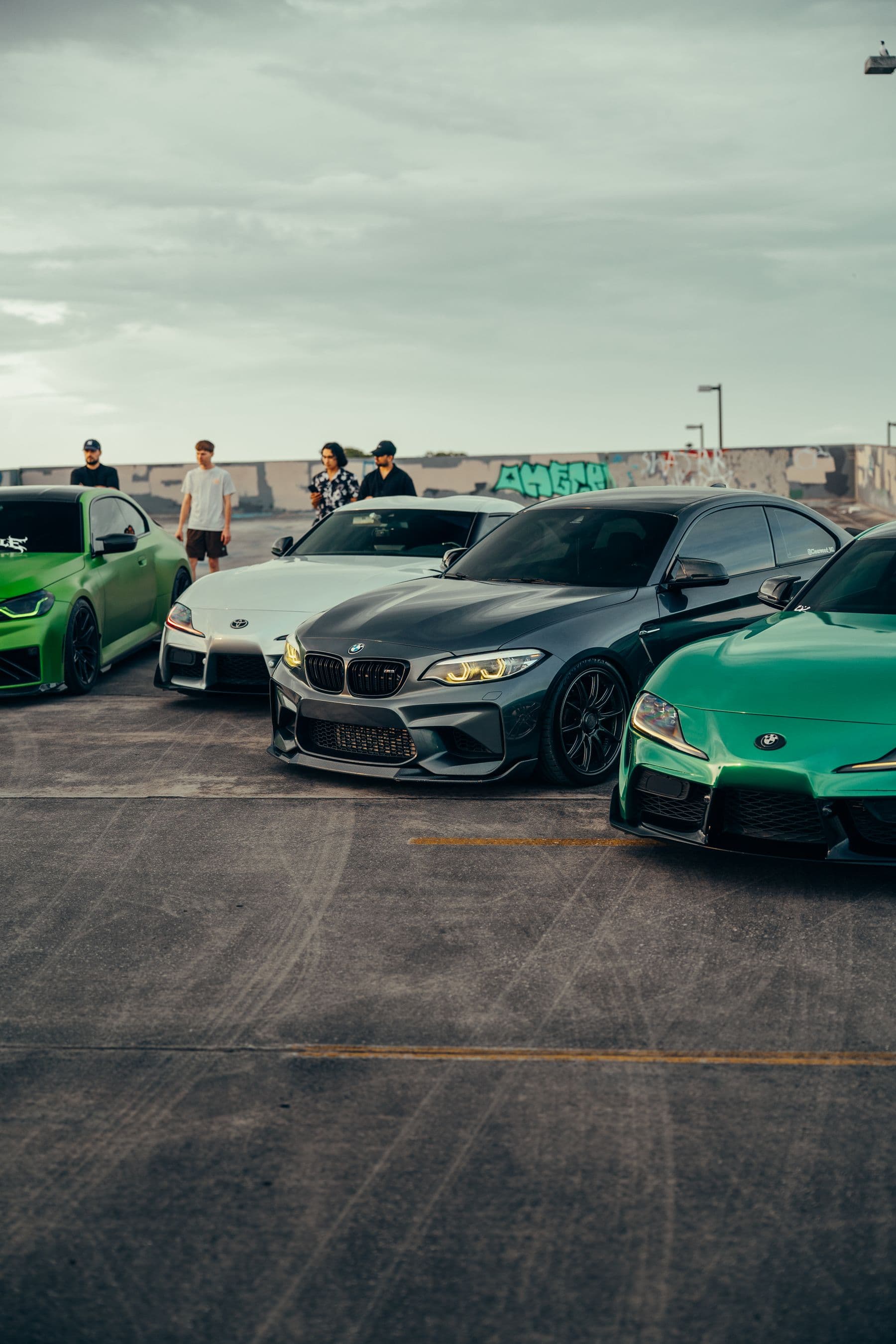 Modified BMWs and Supras lined up at a rooftop meet in front of a graffiti wall